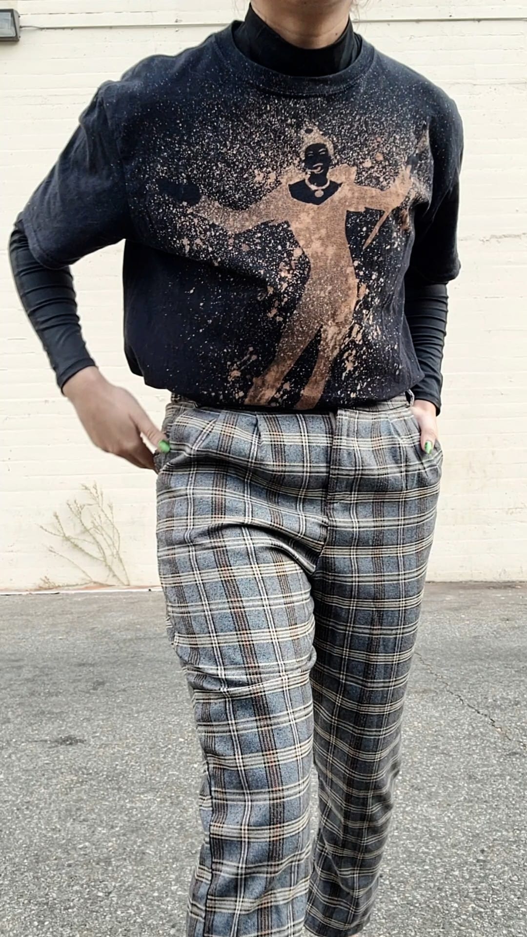 Model posed standing with hand in pocket and wearing black turtleneck cropped with Fifth Element t-shirt; pants worn are a grey plaid.