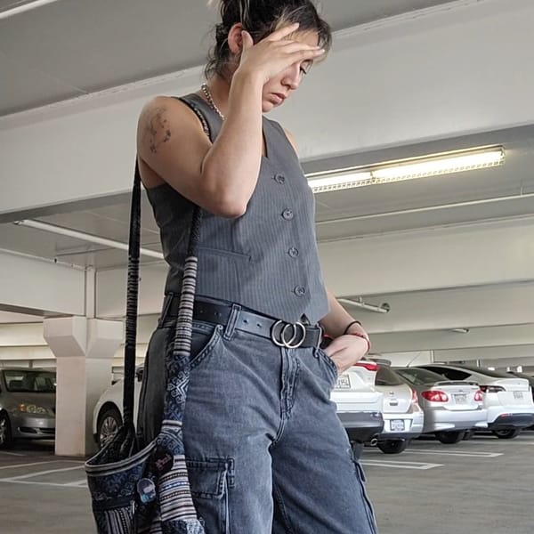 Outfit worn: grey striped vest, dark jeans, silver necklace, black belt, crossbody. Pose: hand covers face in parking garage