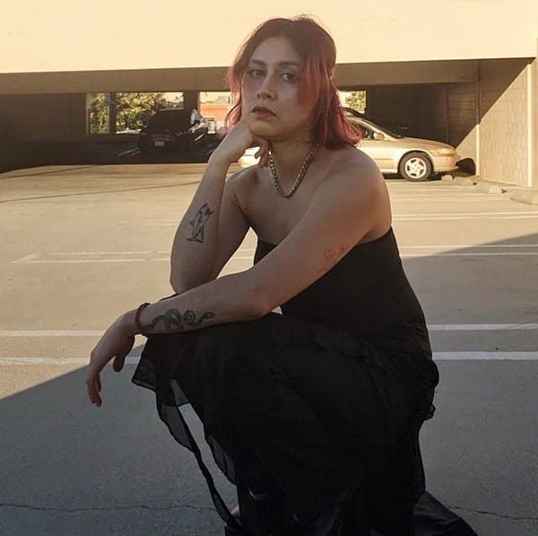 Model in a crouched position wearing a black dress with a long spiral ruffle fringe over black pants and tall boots. 