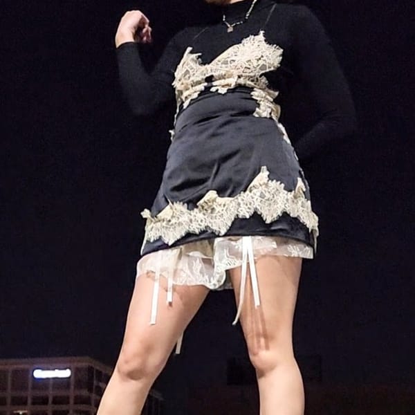 Model posed standing wearing nightgown with lace accents, black turtleneck, and boots. Dress is layered over two skirts.