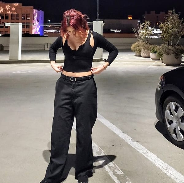 Model poses standing, adjusting shirt. Wears striped pleated pants with black tank top and shrug held together by a buckle.